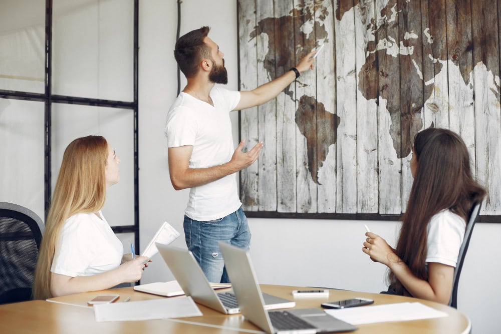 Man points to a world map mural on the wall while two women listen in a meeting room.