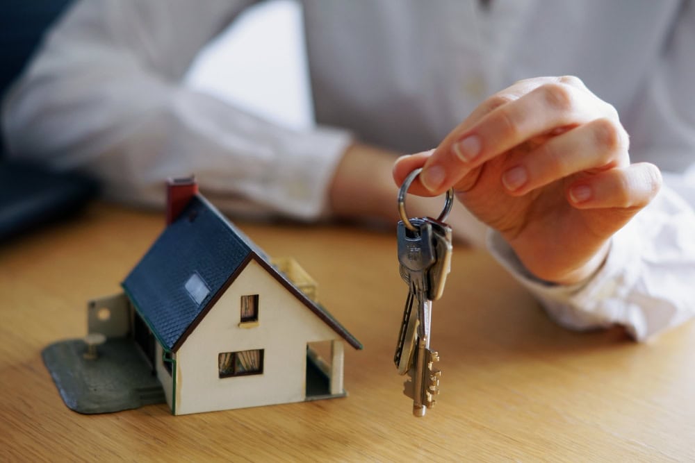 Hand holding a key ring with several keys beside a miniature house model on a wooden table, symbolizing home ownership.