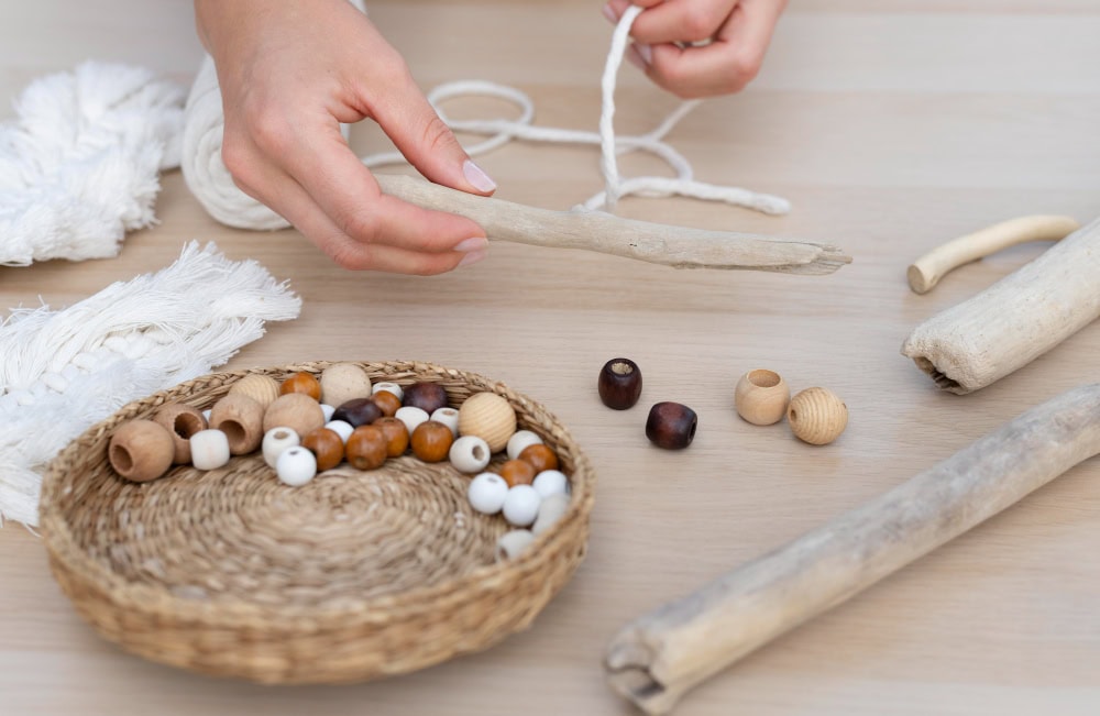 Hands stringing wooden beads with white twine on a craft table; beads in a basket nearby