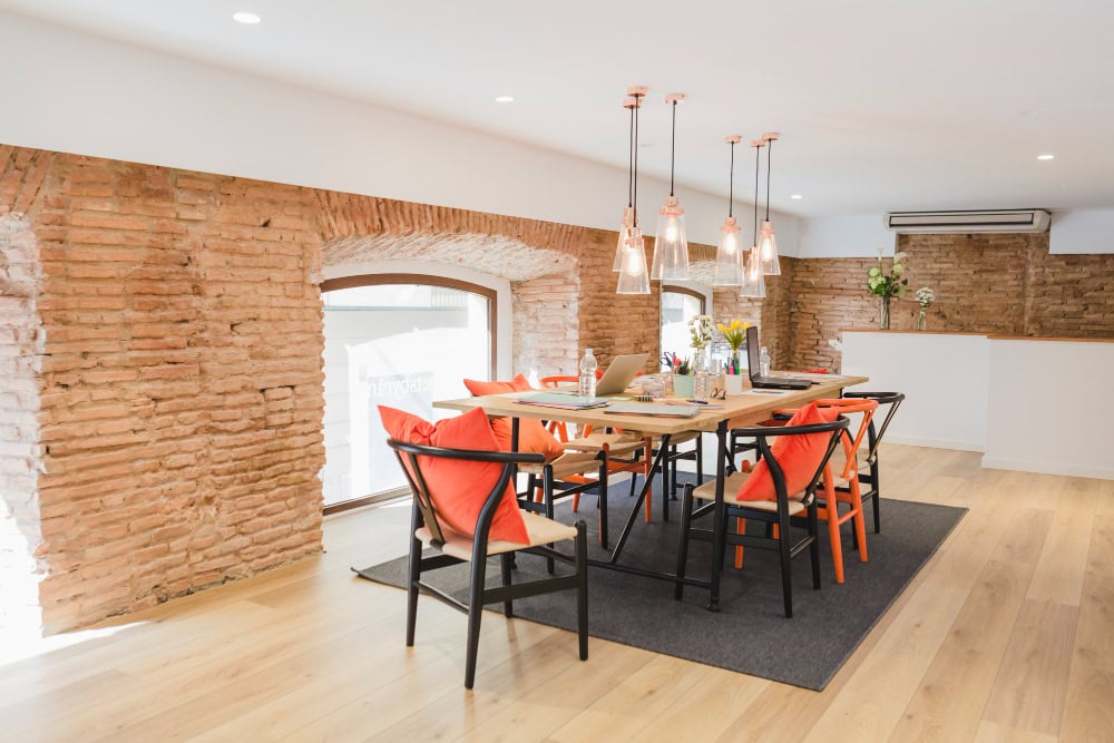 Bright coworking space with a long wooden table, orange-cushioned chairs, and glass pendant lights over exposed brick arches.
