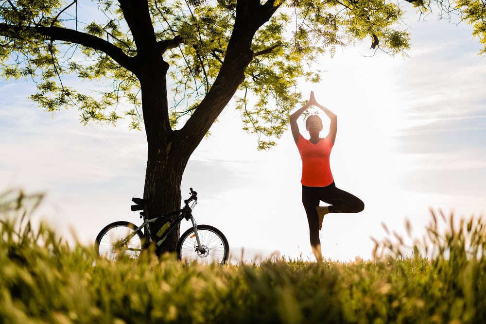 Person doing a tree pose in a sunlit park with a bicycle propped against a tree nearby