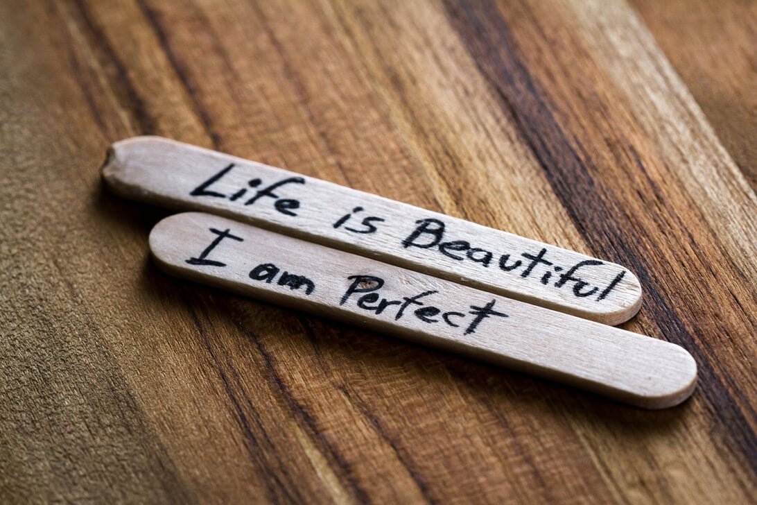Two wooden craft sticks on a wood surface with handwritten phrases: 'Life is Beautiful' and 'I am Perfect'.