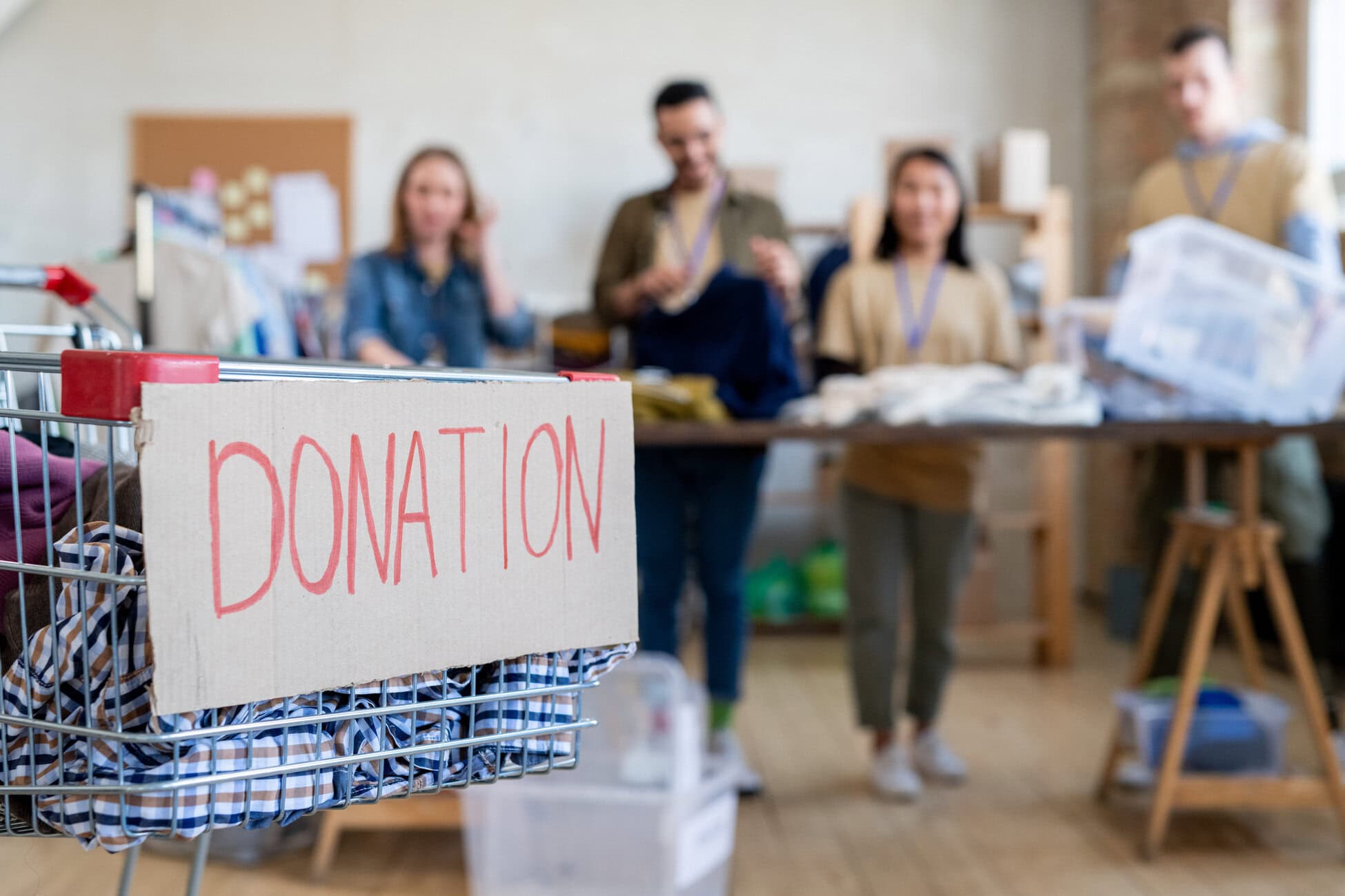 Cart with a cardboard sign reading DONATION in red letters, donation items inside; volunteers in the background organizing supplies in a bright room.