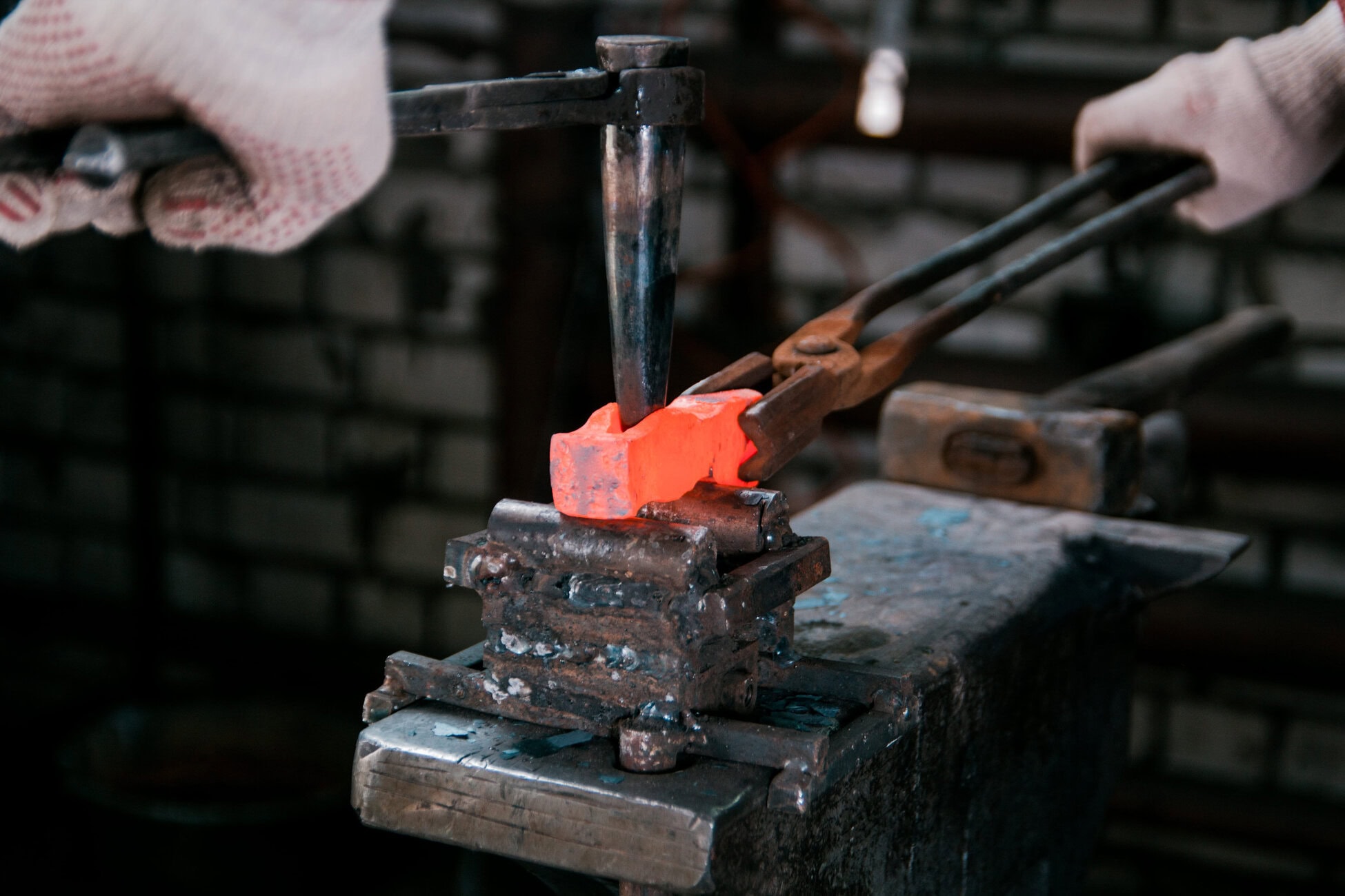 Blacksmith wearing gloves shaping a glowing orange-hot metal with tongs and a hammer over an anvil/vise.