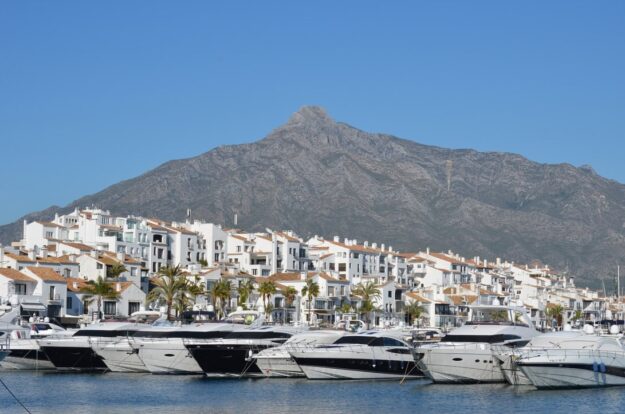 Marina with luxury yachts in a harbor, white Mediterranean buildings, palm trees, and a rocky mountain backdrop.