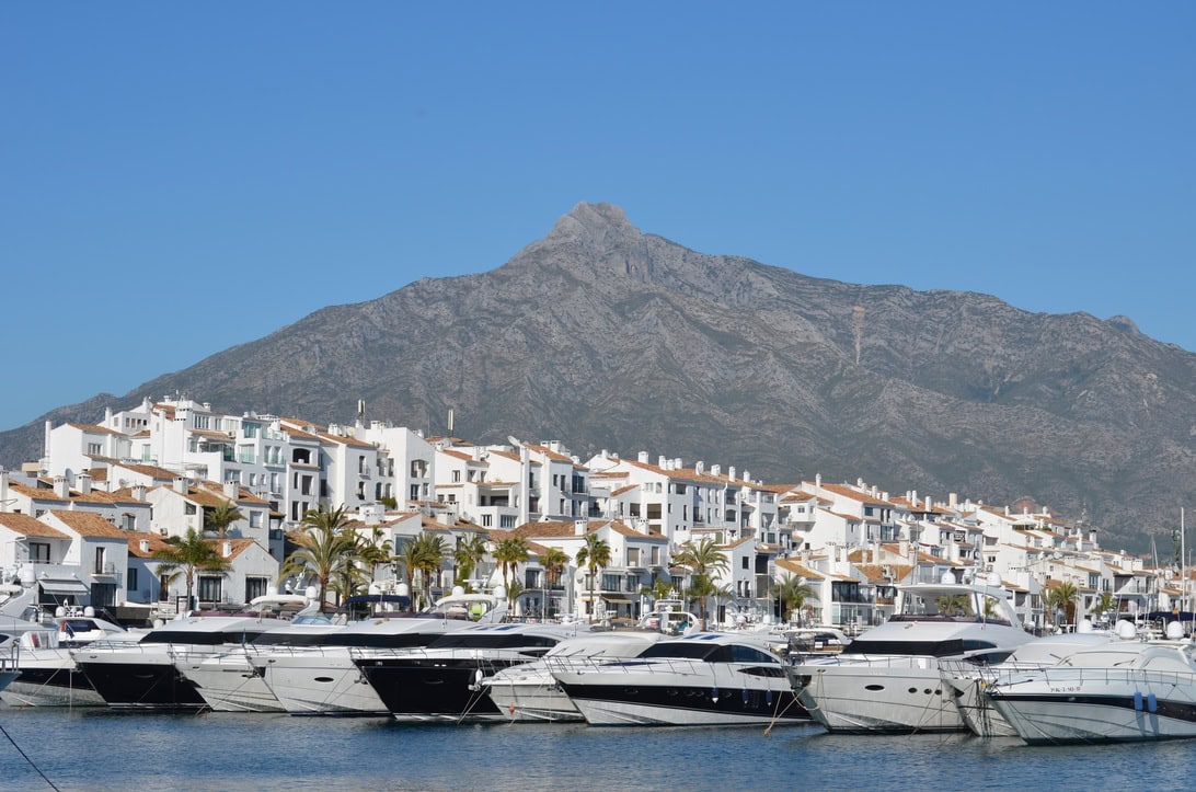 Marina with luxury yachts in a harbor, white Mediterranean buildings, palm trees, and a rocky mountain backdrop.