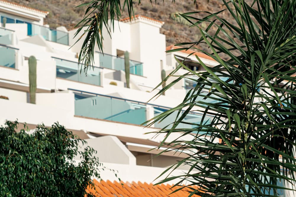 White Mediterranean-style apartment complex with glass balconies seen through palm fronds in the foreground.