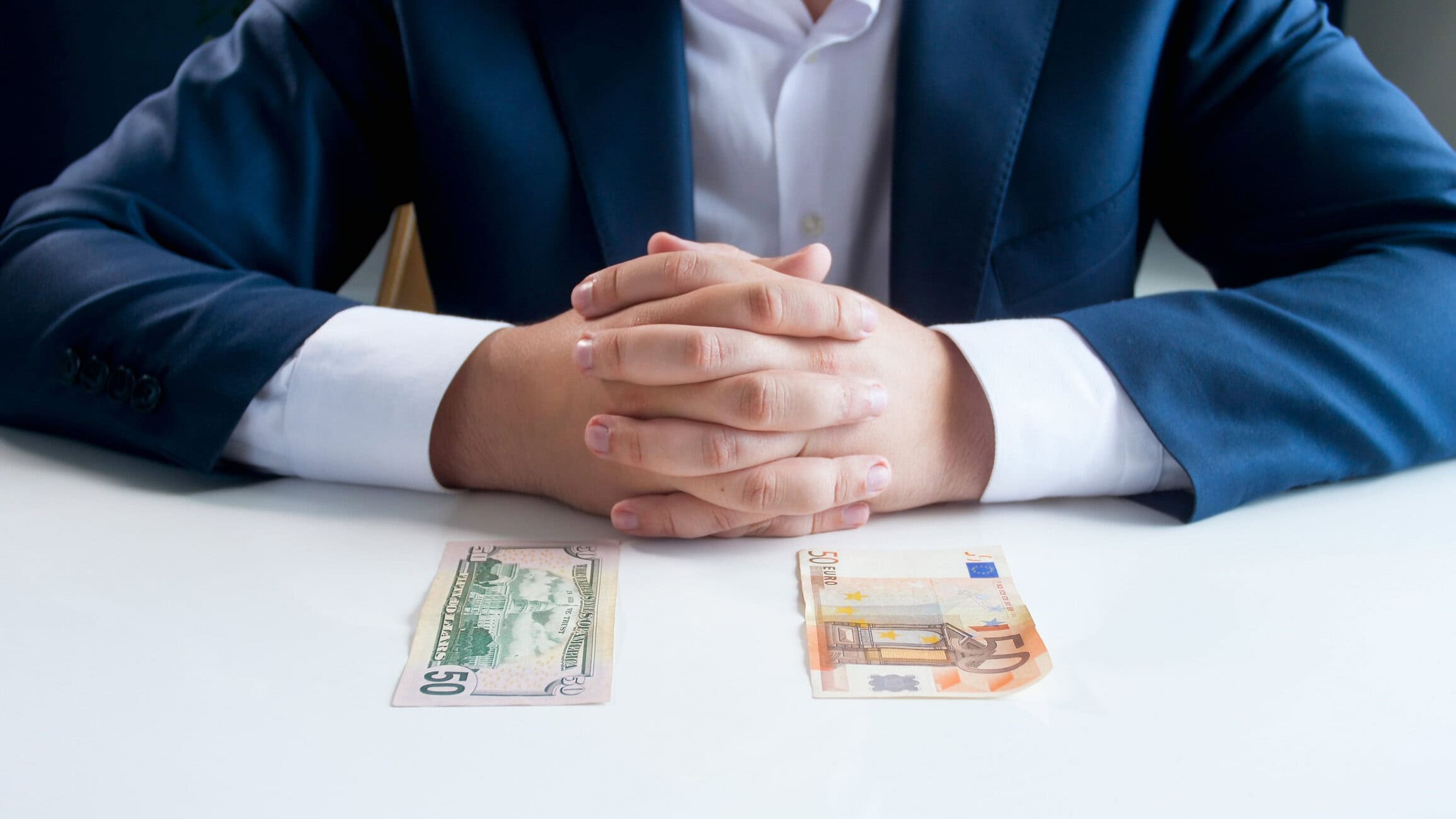 Person in a navy suit sits at a table with hands folded; two euro banknotes (50 and 20 euro) are laid out in front.