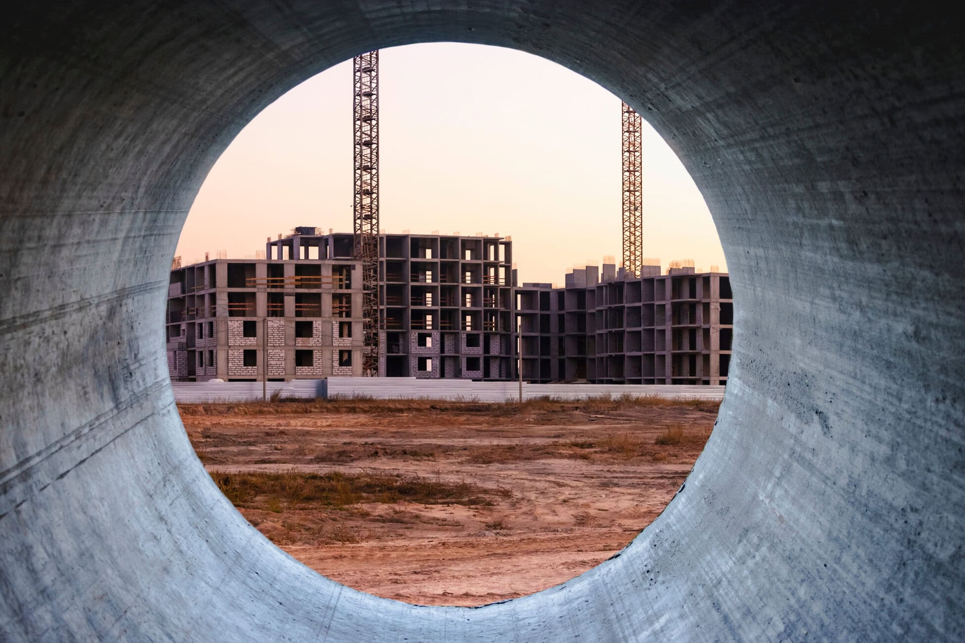 View of a construction site framed by a large concrete pipe, showing unfinished buildings and two cranes at sunset.