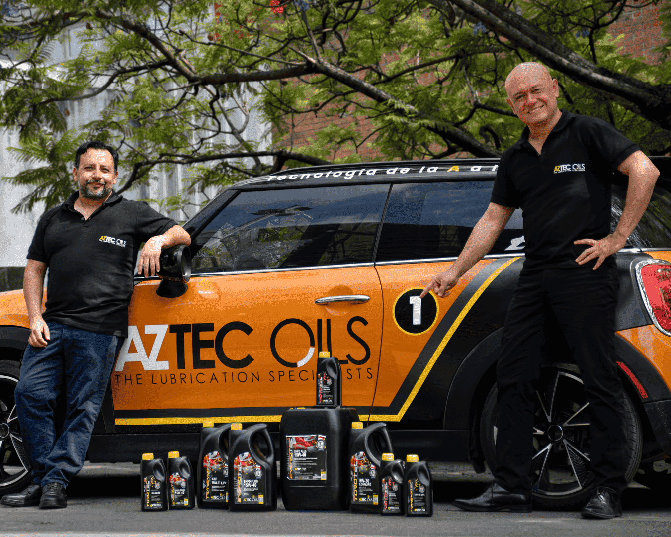 Two men in black shirts stand beside an orange AZTEC OILS car with various lubricant containers arranged in front.