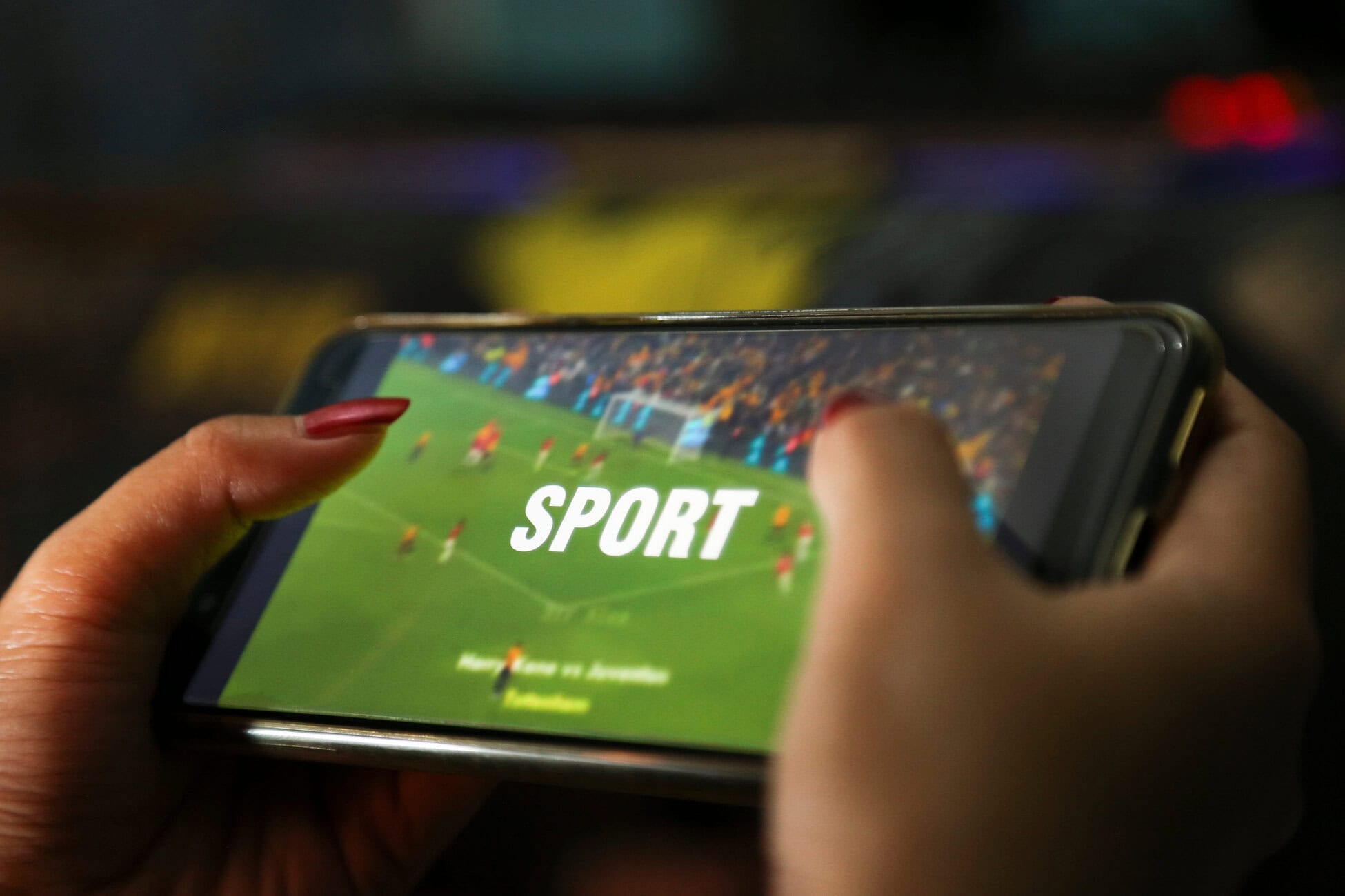 Close-up of hands holding a smartphone while playing a soccer game; the screen shows a green field, players, and the word SPORT in bold white letters.