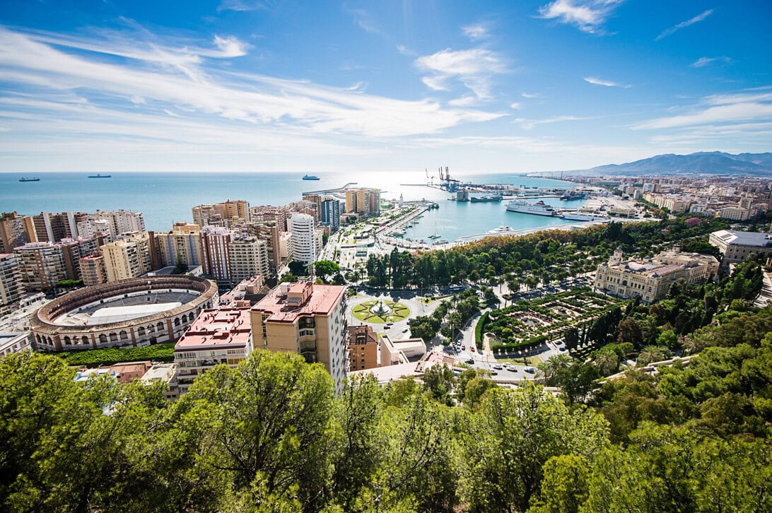 A coastal cityscape with a large circular stadium, dense buildings, a marina, and a bright blue sea under a partly cloudy sky.