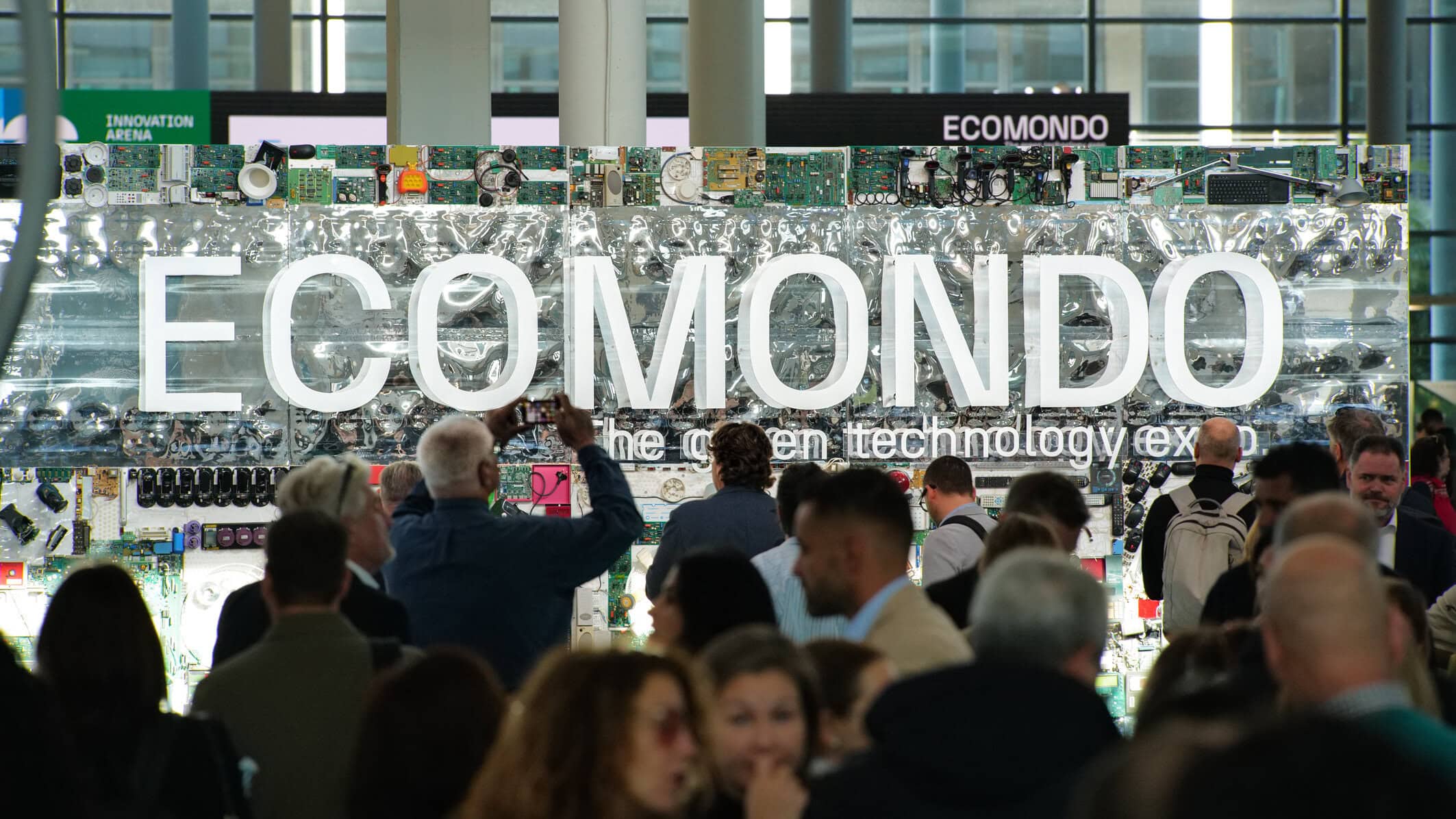 Crowd of people at a technology expo in front of a wall covered with circuit boards and a large 'ECOMONDO' sign.