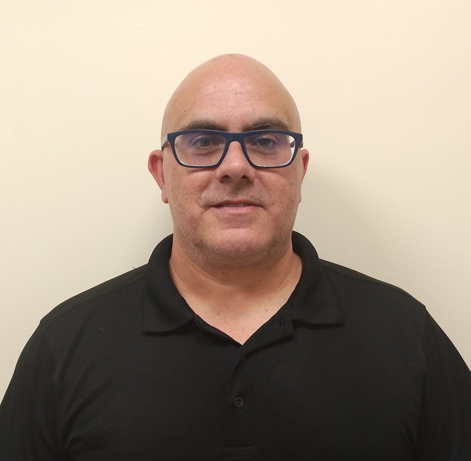Close-up of a bald man wearing blue-framed glasses and a black polo shirt against a light beige wall.
