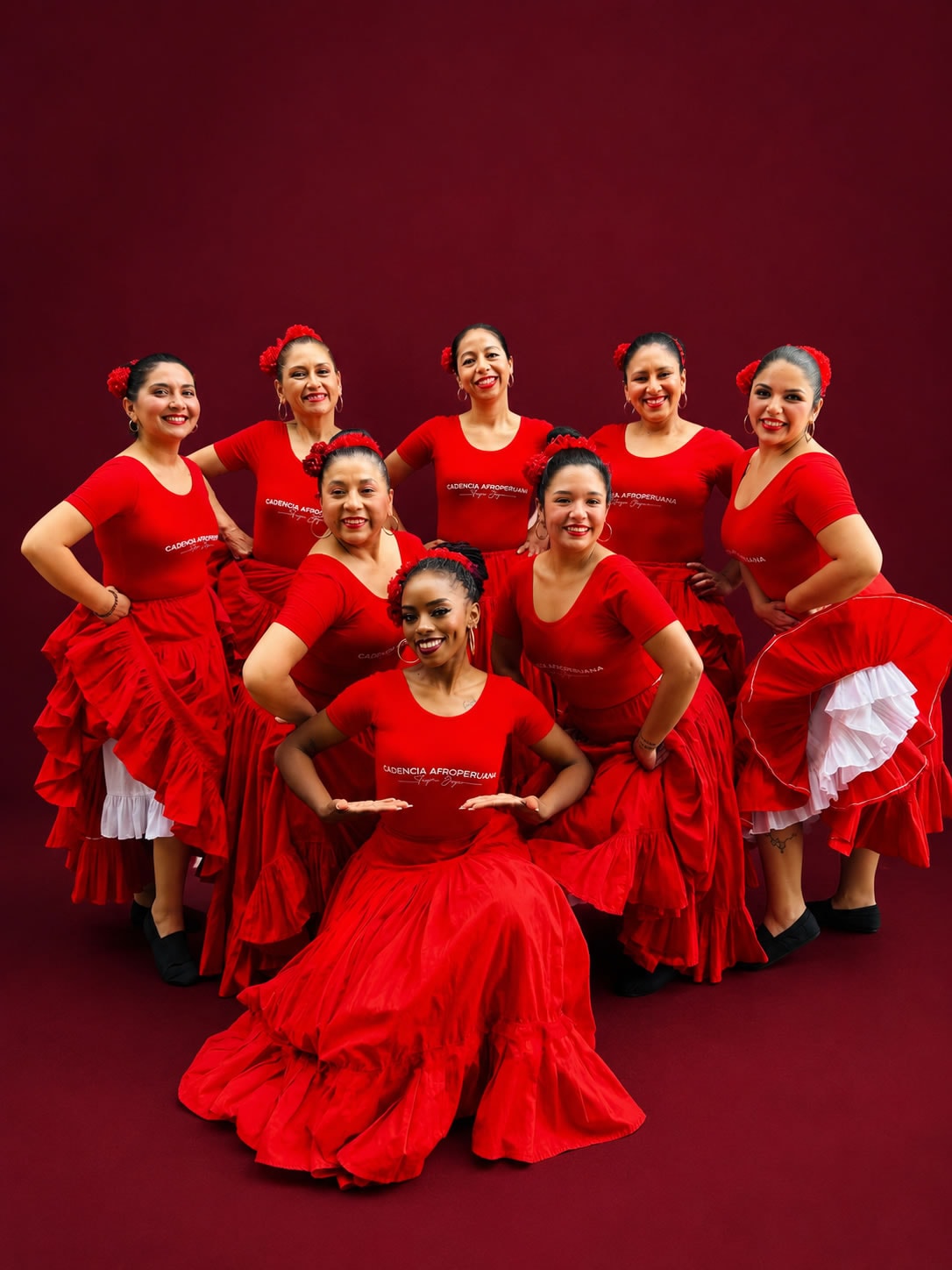 Afro-Peruvian dance group of nine women in red dresses posing together against a red backdrop with smiles.