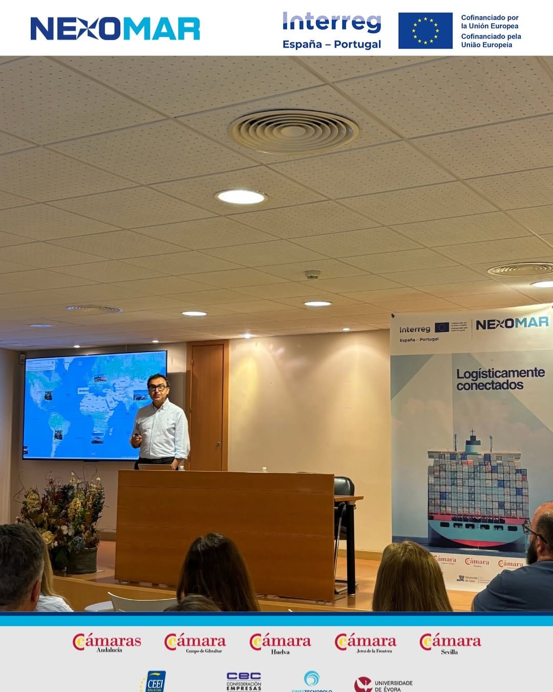 Male presenter at a wooden podium in a conference room, with a world map on screen behind him; banners for Interreg/NEXOMAR and a cargo-ship graphic flank the scene.