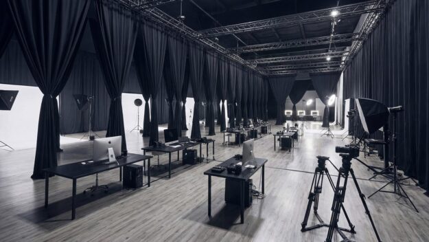 Long row of editing workstations in a professional studio, with lighting rigs and tripods nearby.