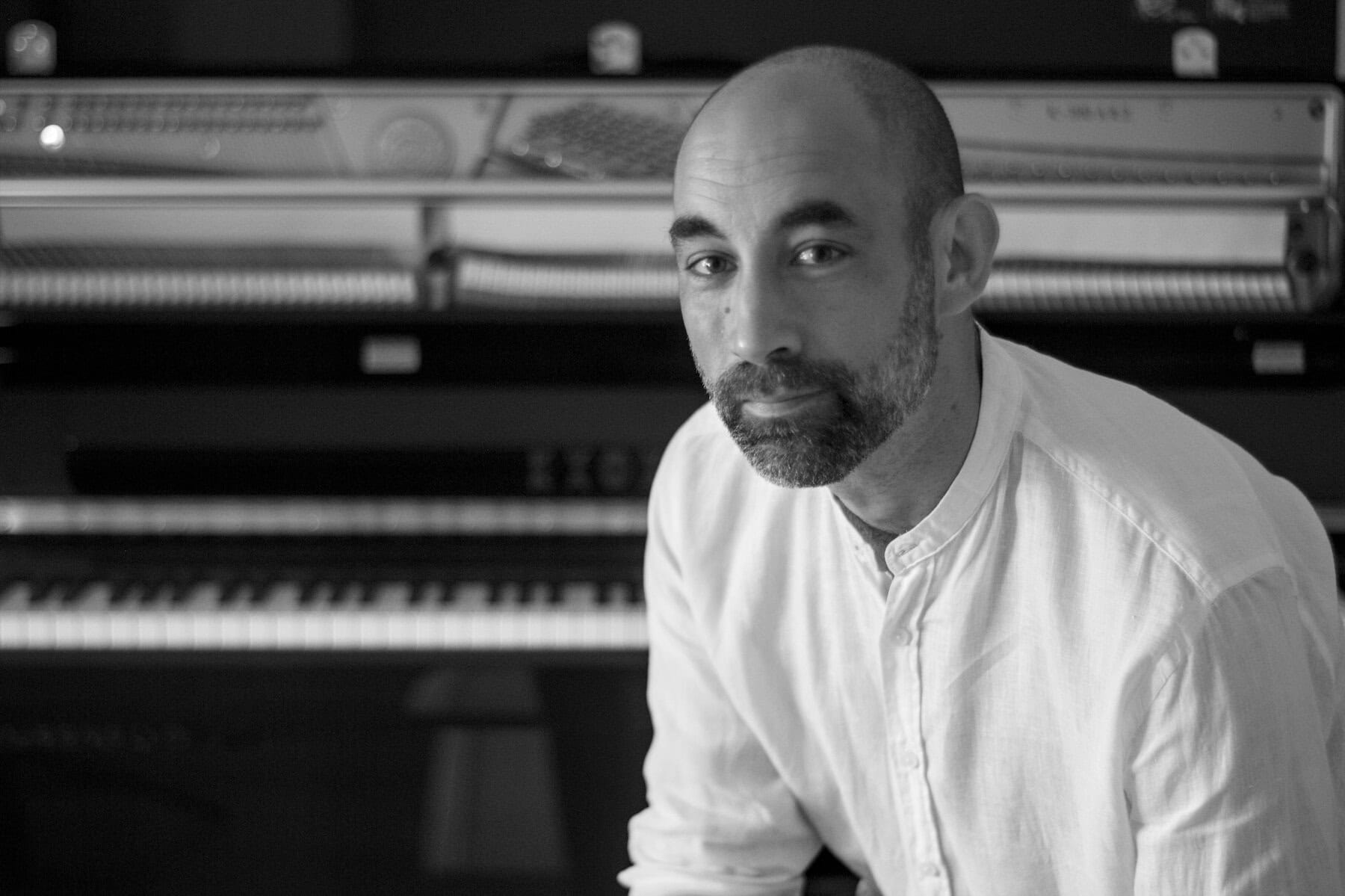 Portrait of a man with a beard in a white shirt, seated beside a piano and looking at the camera (black and white).