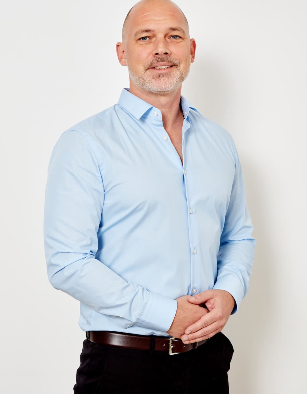 Man in a light blue dress shirt and black belt stands against a white background.