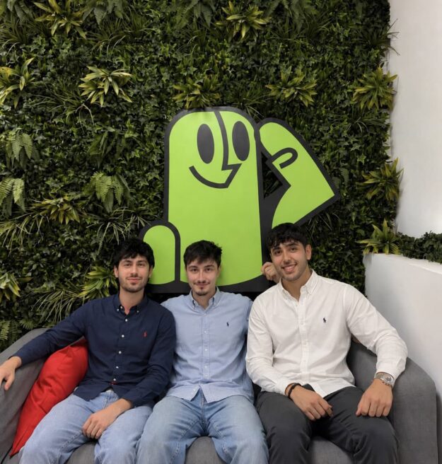 Three young men sit on a gray couch, smiling, with a large lime-green cartoon figure on a leafy wall behind them.