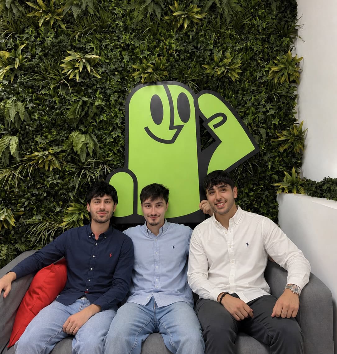 Three young men sit on a gray couch, smiling, with a large lime-green cartoon figure on a leafy wall behind them.