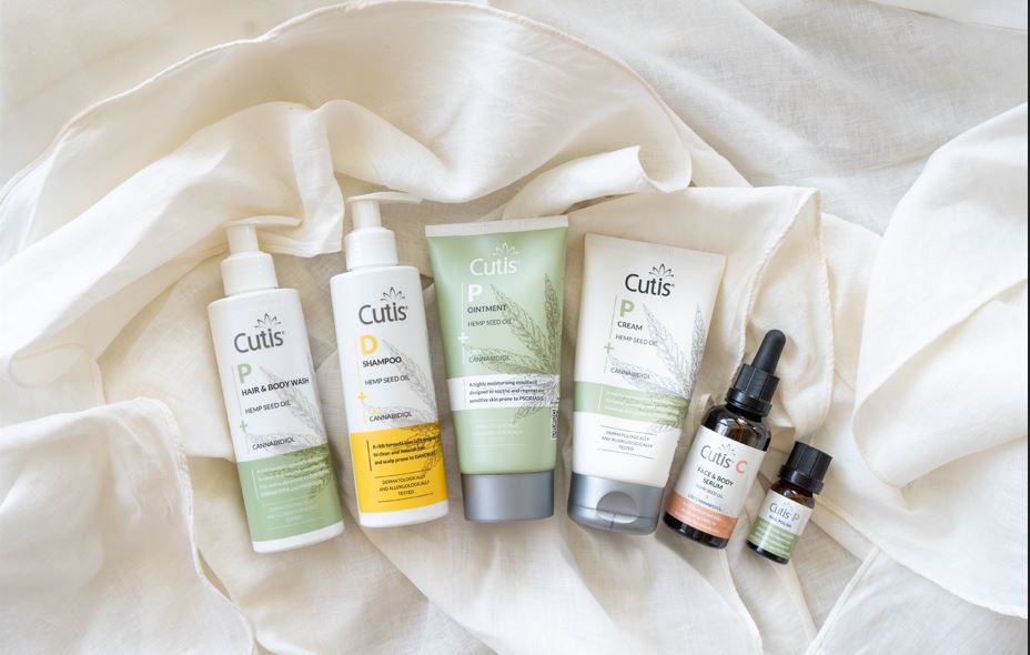 Row of Cutis hemp seed oil skincare bottles and tubes on white fabric, showing shampoo, conditioner/ointment, cream, and oils.