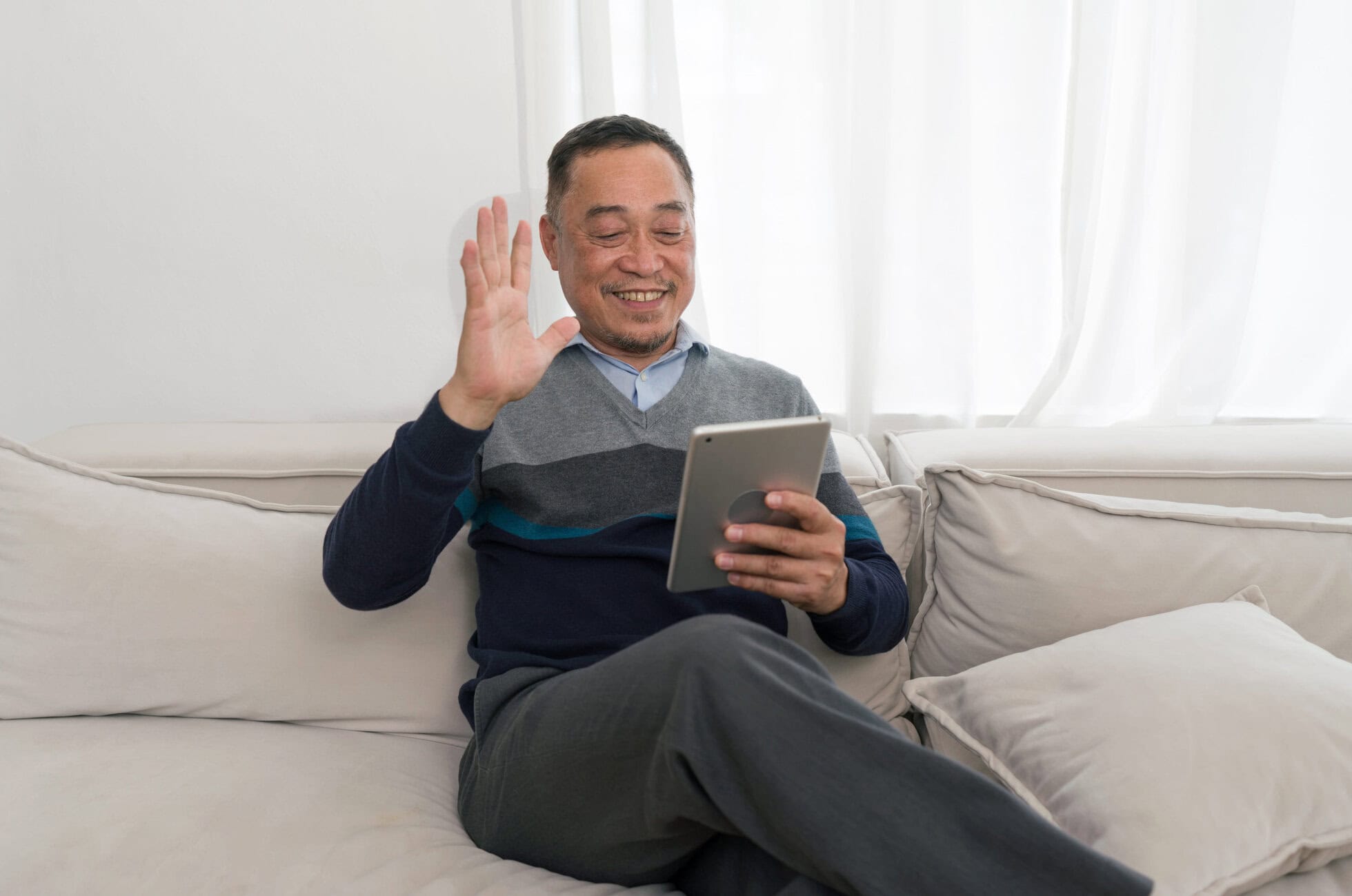 Smiling man on a sofa waving at a tablet he’s holding. Living room setting with white curtains in the background.
