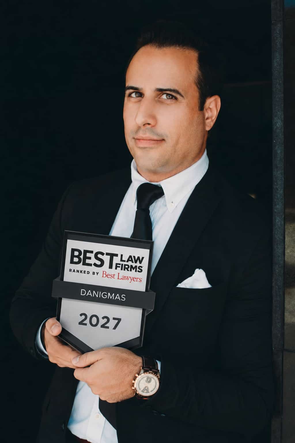 Portrait of a man in a black suit and white shirt holding a 'Best Law Firms 2027' award with the name 'DANIGMAS' on the plaque.