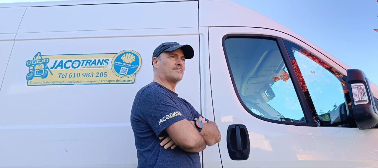 Man in a blue t-shirt and cap stands with arms crossed beside a white Jacotrans delivery van with logo on the side.