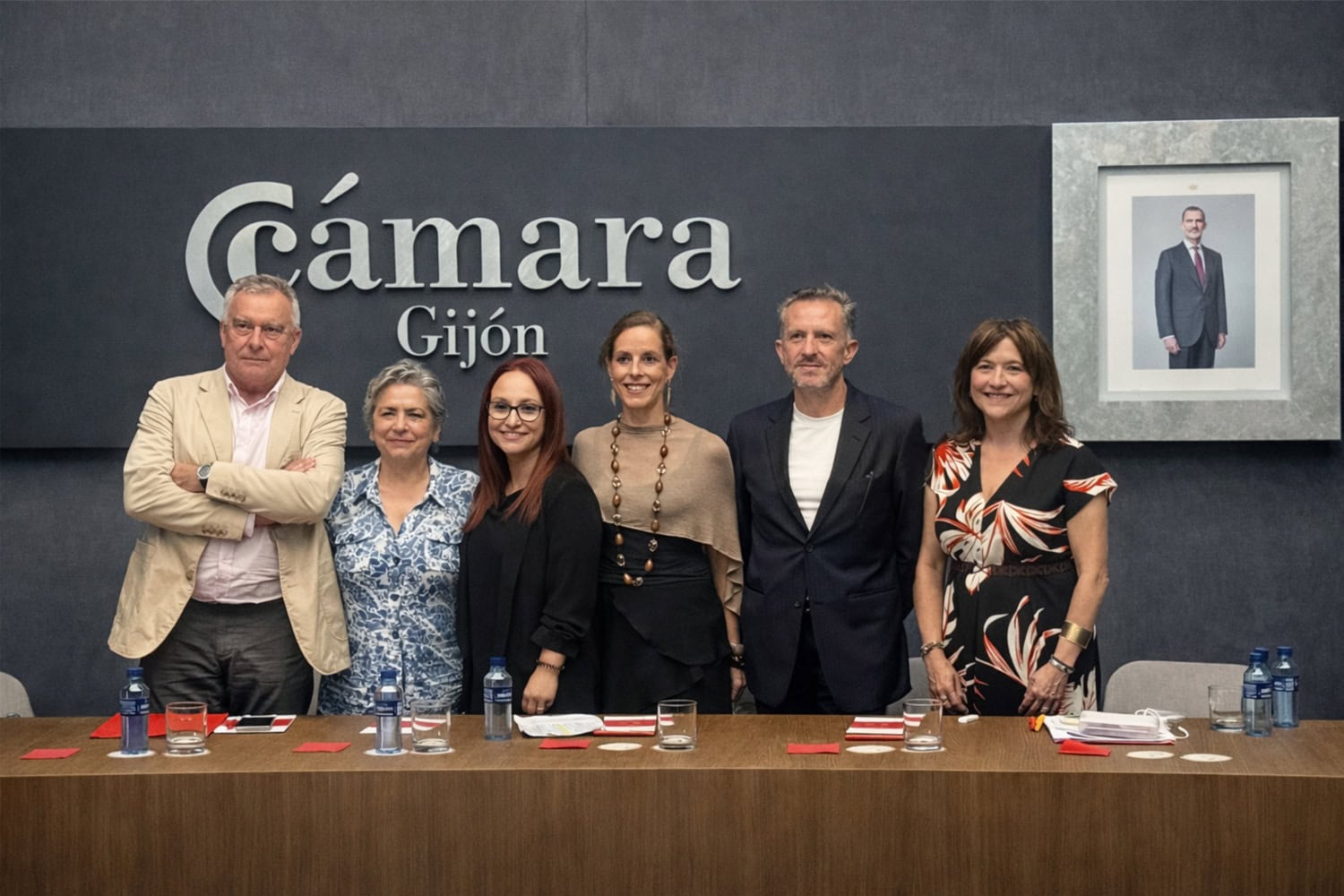 Six adults stand shoulder to shoulder behind a conference table in a room with a dark wall and a large 'Cámara Gijón' sign; water bottles and red notepads on the table.