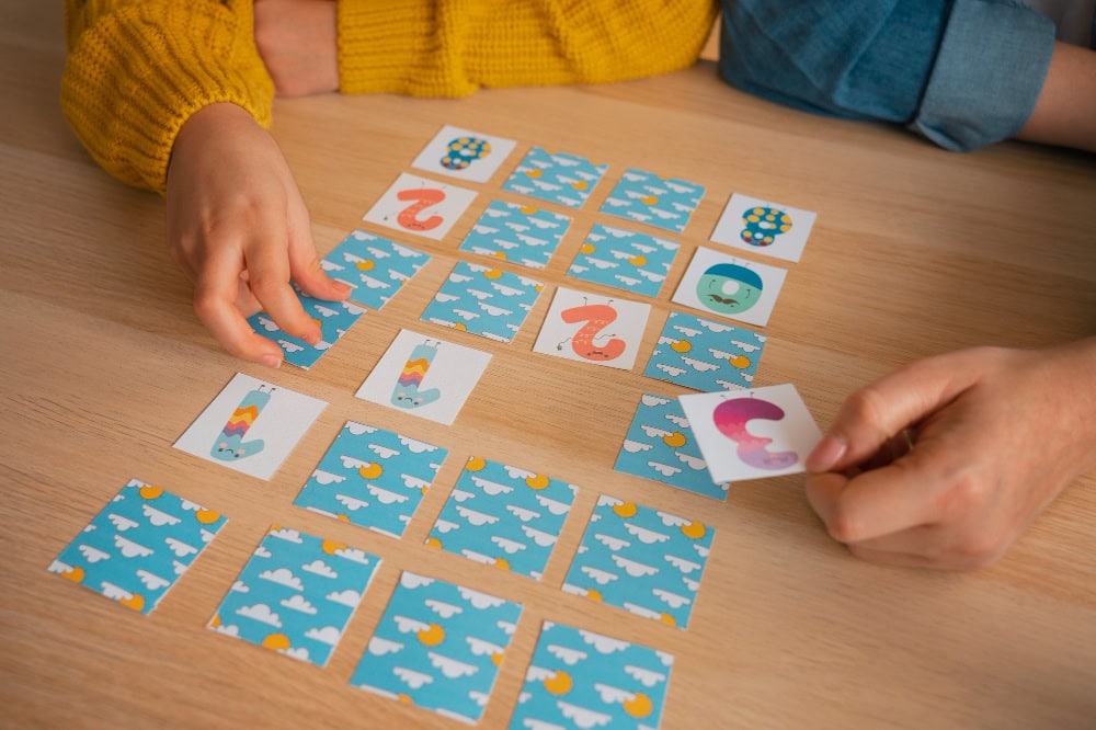 Two people play a card-matching game on a wooden table, arranging blue-pattern back cards with a few face-up tiles showing numbers and shapes on top.