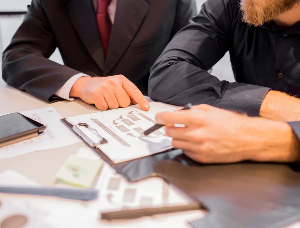 Two colleagues in suits review a document on a clipboard at a desk, with a smartphone nearby.