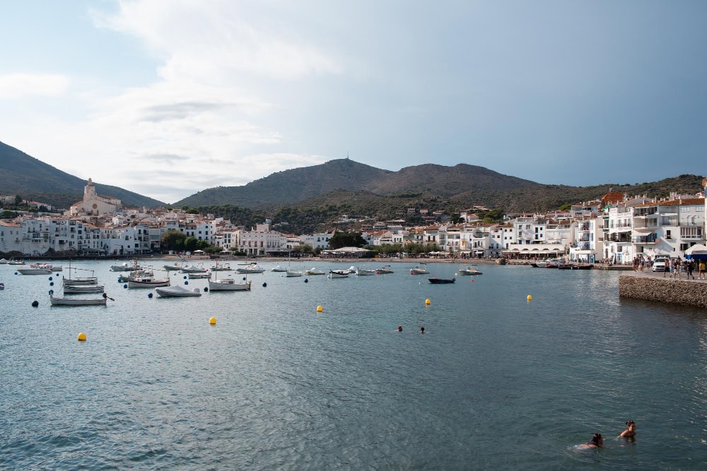 Harbor town with white buildings along the shore, small boats on calm water, hills in the distance.