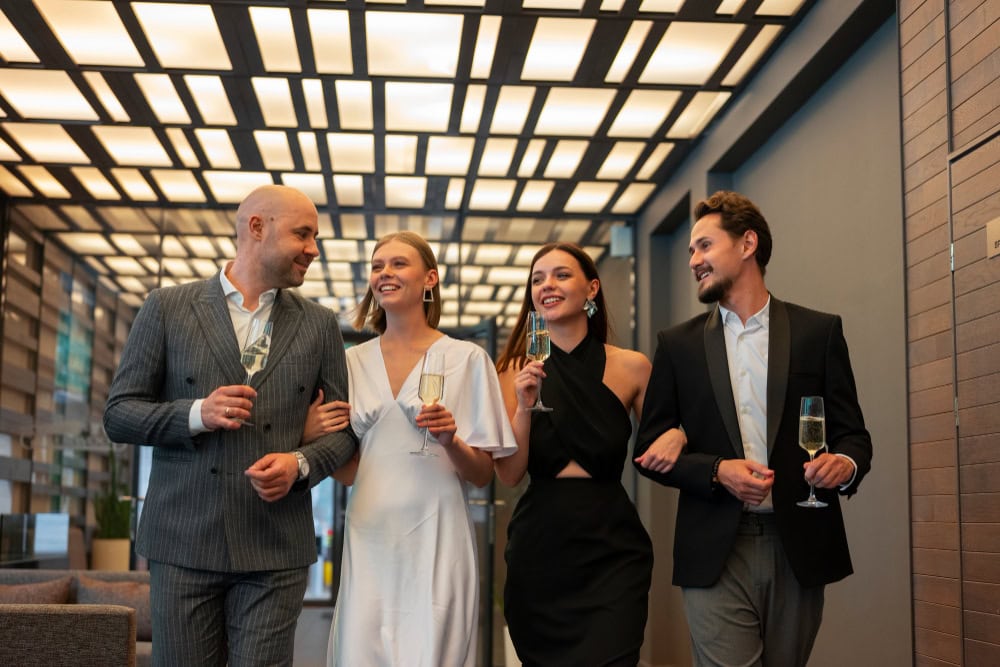 Four professionally dressed adults walk arm-in-arm, smiling and holding champagne flutes in a modern hallway recption setting.