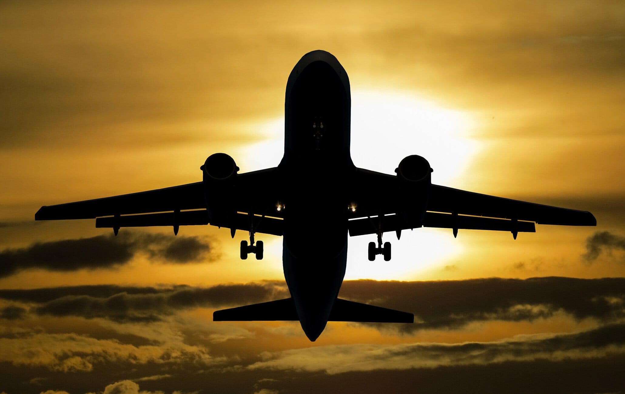 Front-on silhouette of a large passenger jet with landing gear down, descending toward a golden sunset.
