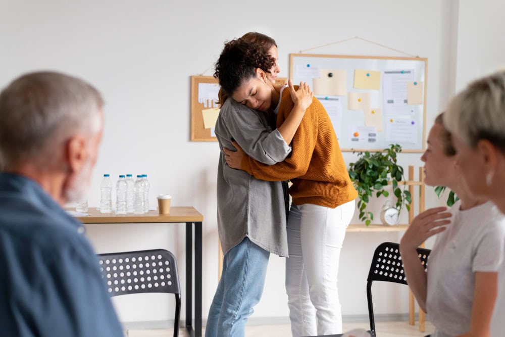 Two people hug in a bright room during a group therapy or support meeting, with others seated in the background.