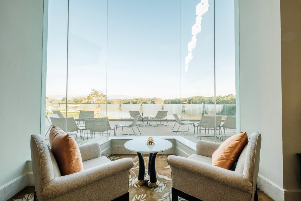 Two beige armchairs with orange cushions flank a small round table in a bright seating area by a large glass window overlooking an outdoor patio and lake.