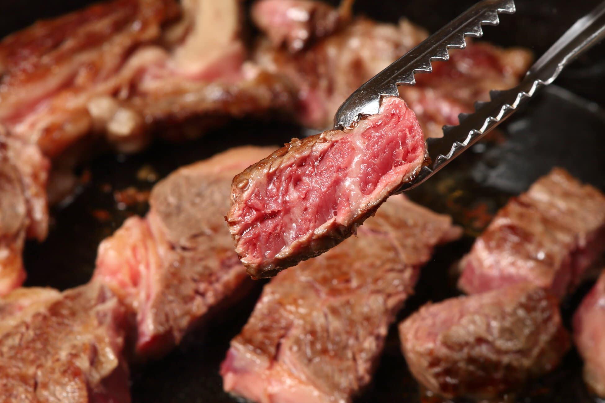 Close-up of a pink, juicy beef rib piece being lifted with metal tongs over a grill.