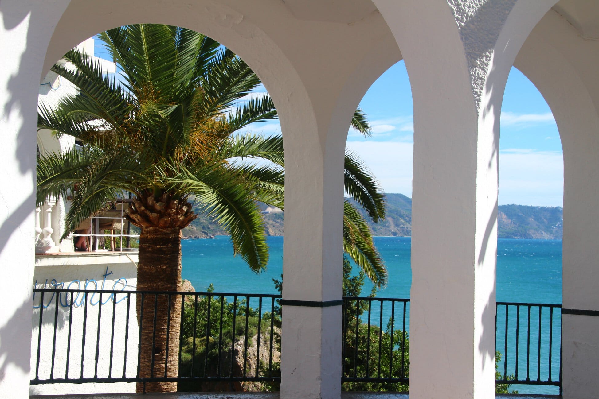White arches frame a seaside view: palm trees, blue sea, and distant hills beyond a black railing.