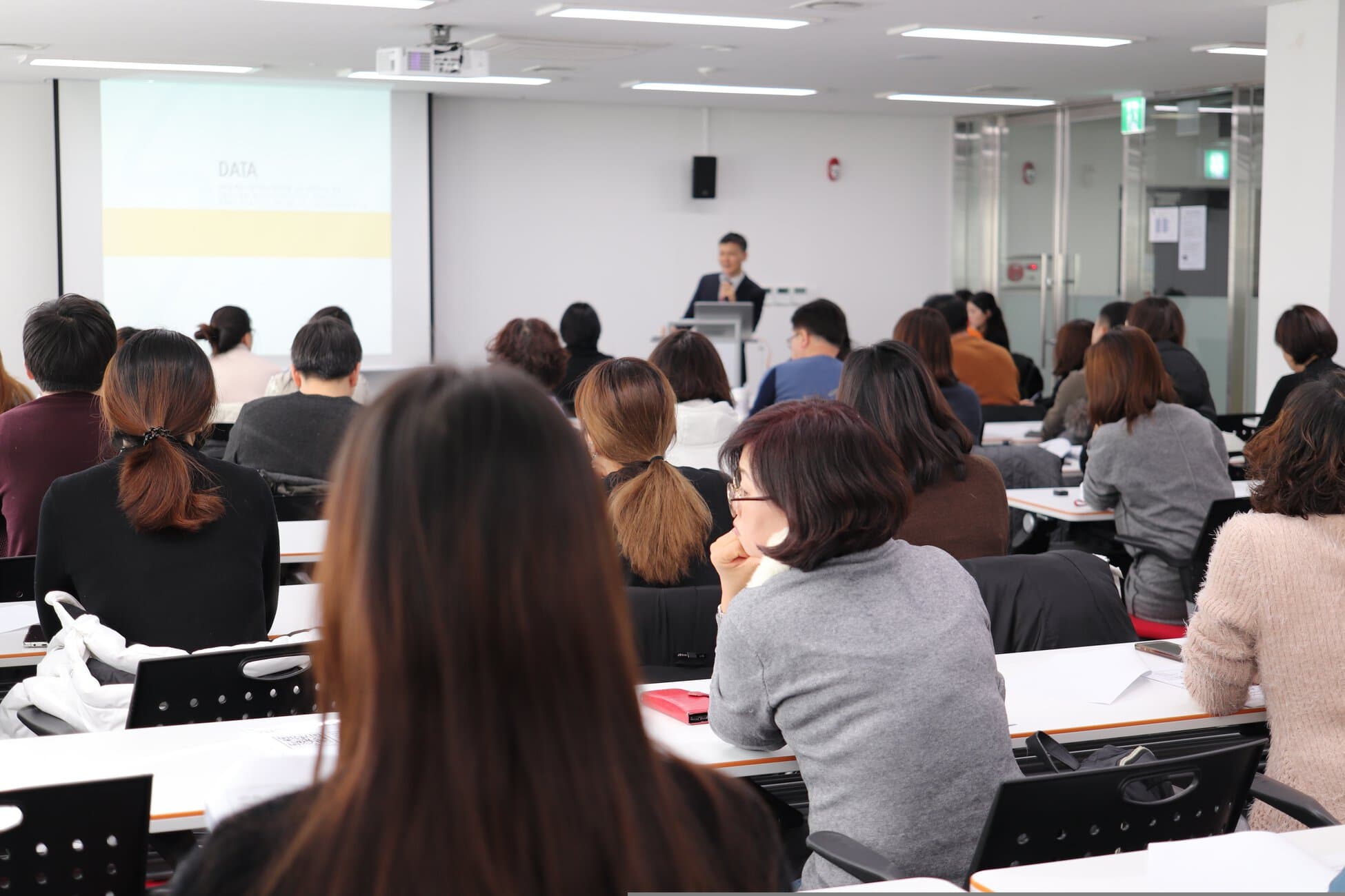 Audience seated in a lecture hall watching a presenter at the front near a projector screen.