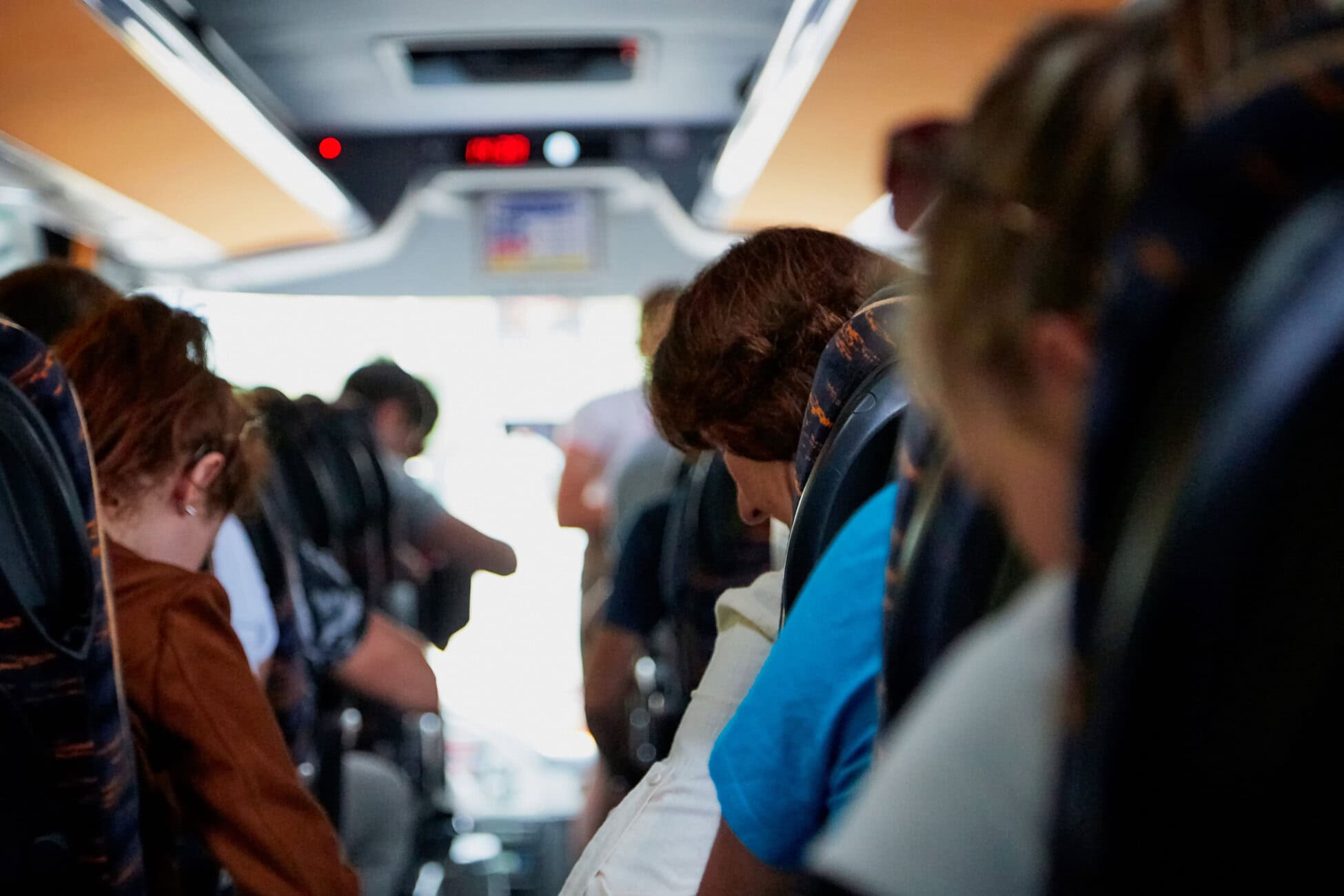 Passengers seated on both sides of a bus aisle, viewed from behind, with a bright rear exit and head tilting forward in the foreground.