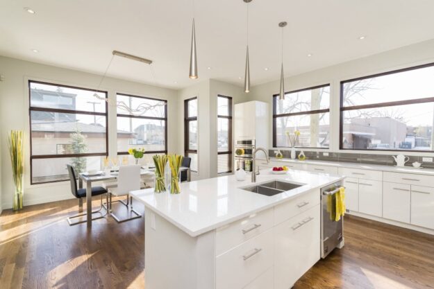 Bright, modern kitchen with white cabinetry, quartz island, and triple pendant lights by a sunny dining area in front of large windows.