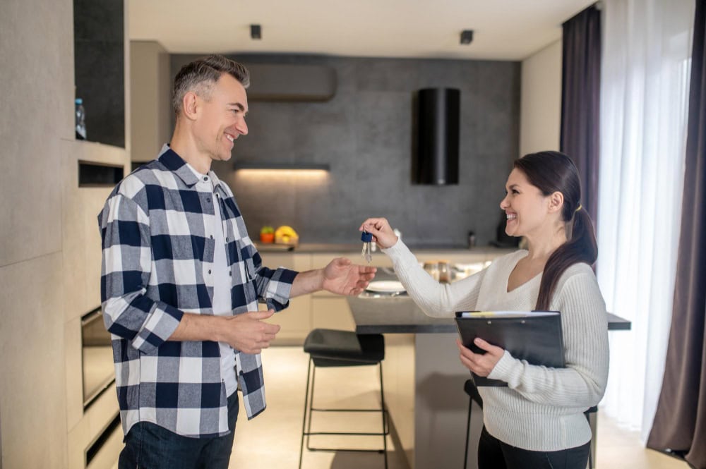 Man in a plaid shirt hands over house keys to a smiling woman in a modern kitchen; she holds a tablet and a folder.