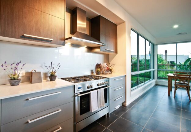 Modern kitchen with stainless steel range, hood, and dark wood cabinets along a white countertop. A bright dining nook and large windows outside.