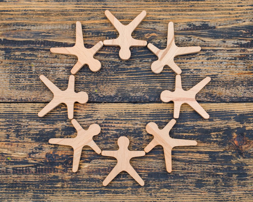 Eight light wood human-shaped figures arranged in a circle on a dark, weathered wooden surface, forming a ring.