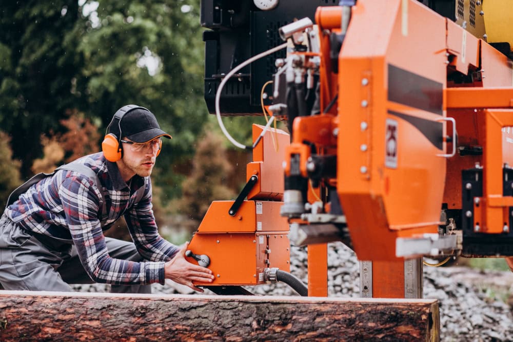 Worker wearing safety earmuffs and cap operates an orange lumber-processing machine outdoors beside a log on a construction site
