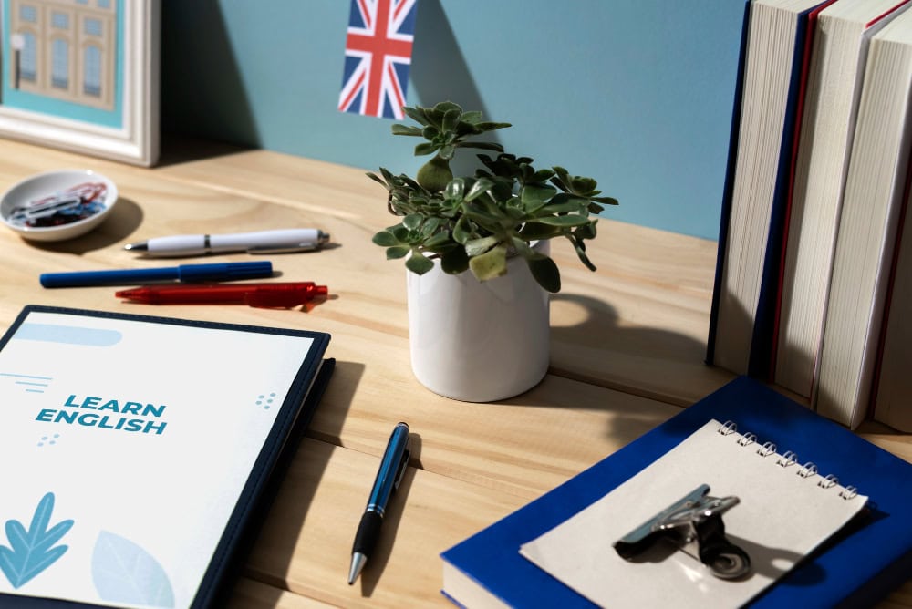A tidy study desk for learning English, with a 'Learn English' workbook, pens, a small plant, a Union Jack flag, and books on a wooden surface.