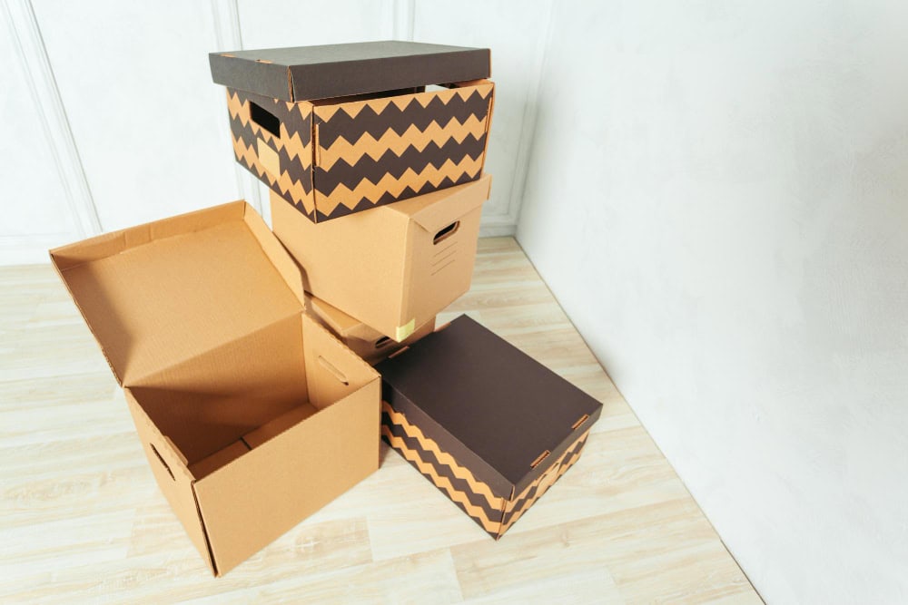 Stack of cardboard moving boxes, some with black-and-brown zigzag patterns, stacked in a corner of a room