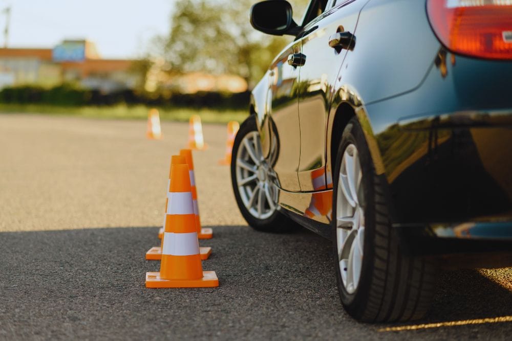 Blue car navigating around orange traffic cones in a parking lot, rear view showing tire and side panel reflections