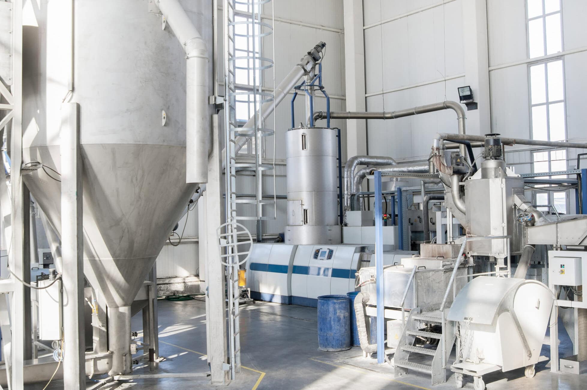 Industrial facility interior with large stainless steel tanks, pipes, and blue support frames.